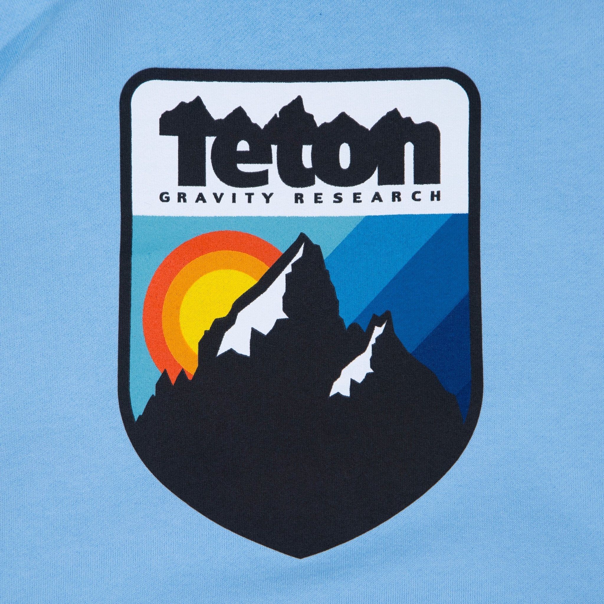 Picture of the logo on the front of the sweatshirt. Shoes the Teton Gravity Research logo with the Grand Tetons and a sunset in the background. #color_baby blue