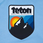 Picture of the logo on the front of the sweatshirt. Shoes the Teton Gravity Research logo with the Grand Tetons and a sunset in the background. #color_baby blue